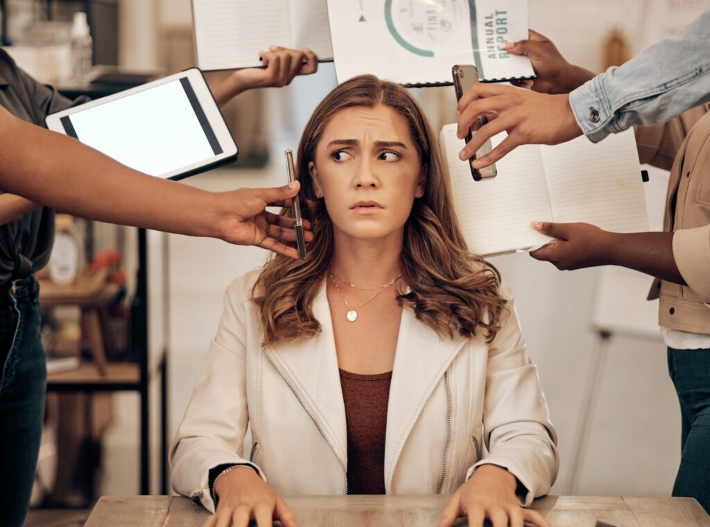 Image Of Woman At Desk With Many Hands Reaching In With Phones, Paperwork, Etc.